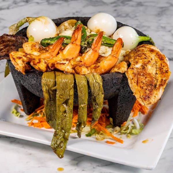 A molcajete filled with grilled shrimp, chicken, steak, onions, and green peppers on a white plate.