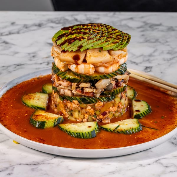 Tall seafood tower with avocado, shrimp, and cucumber, surrounded by red sauce and cucumber slices.