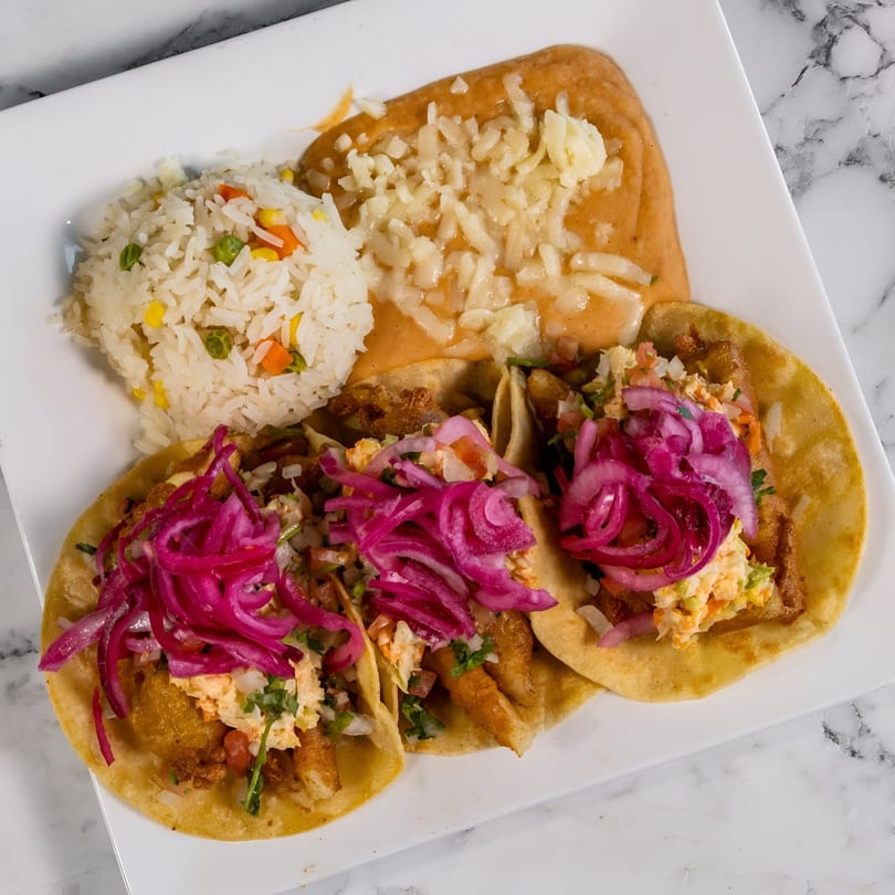 Three Mexican tacos with pickled red onions, rice, and refried beans with cheese on a white plate.